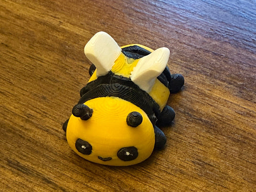 Yellow and black striped 3D printed Mini Flexi Bumblebee with white wings and a kawaii face on a wood table