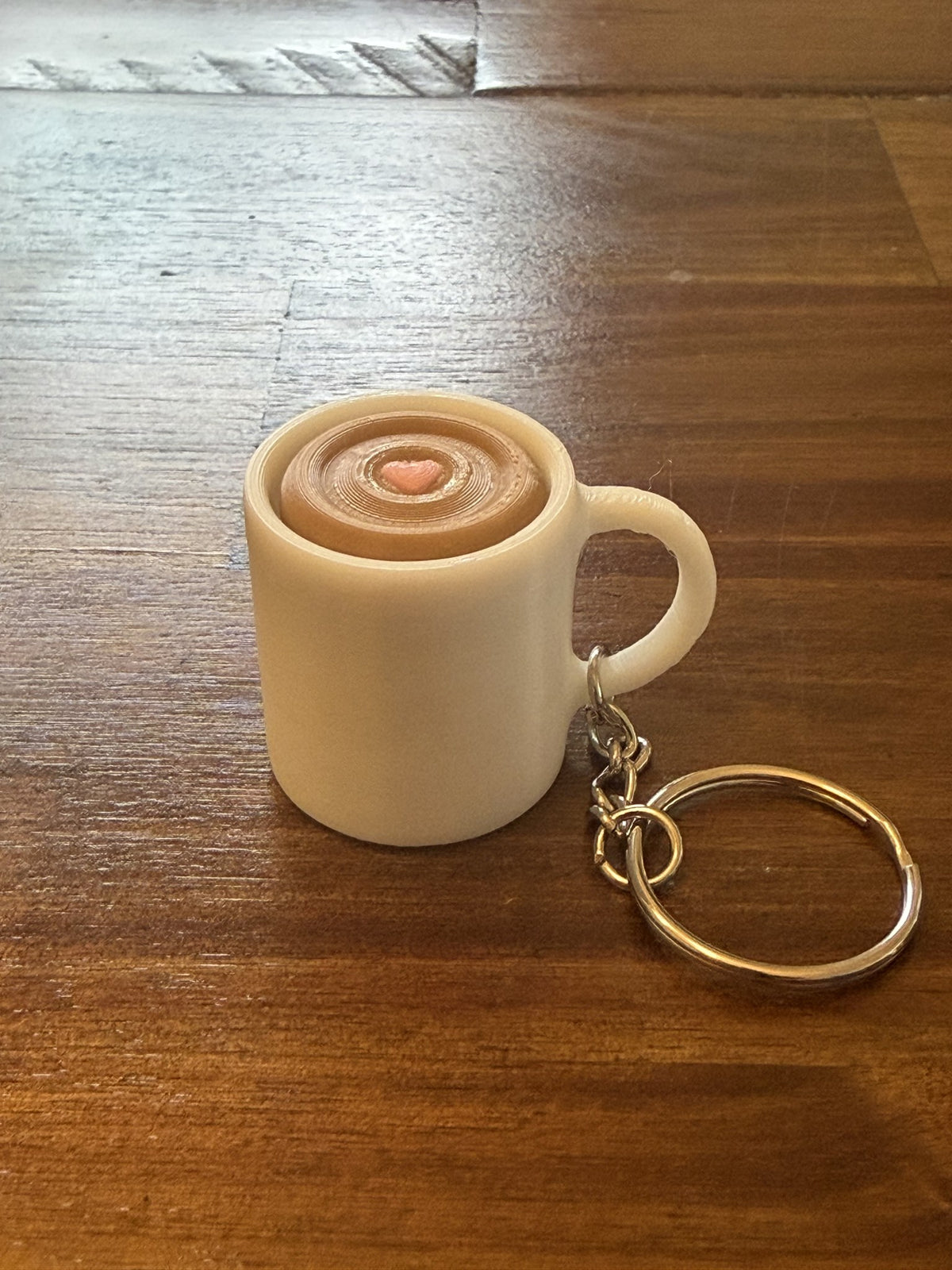 Cream 3D printed Coffee Cup Clicker Keychain shaped like a latte mug with a heart in the foam, on a wood table with its keyring