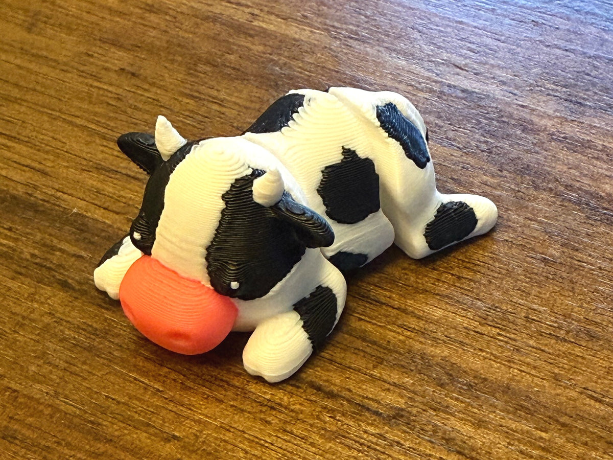 White 3D printed Mini Flexi Cow with black patches and a pink muzzle, three-quarter front view on a wood table