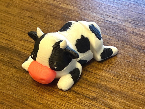 White 3D printed Mini Flexi Cow with black patches and a pink muzzle, three-quarter front view on a wood table