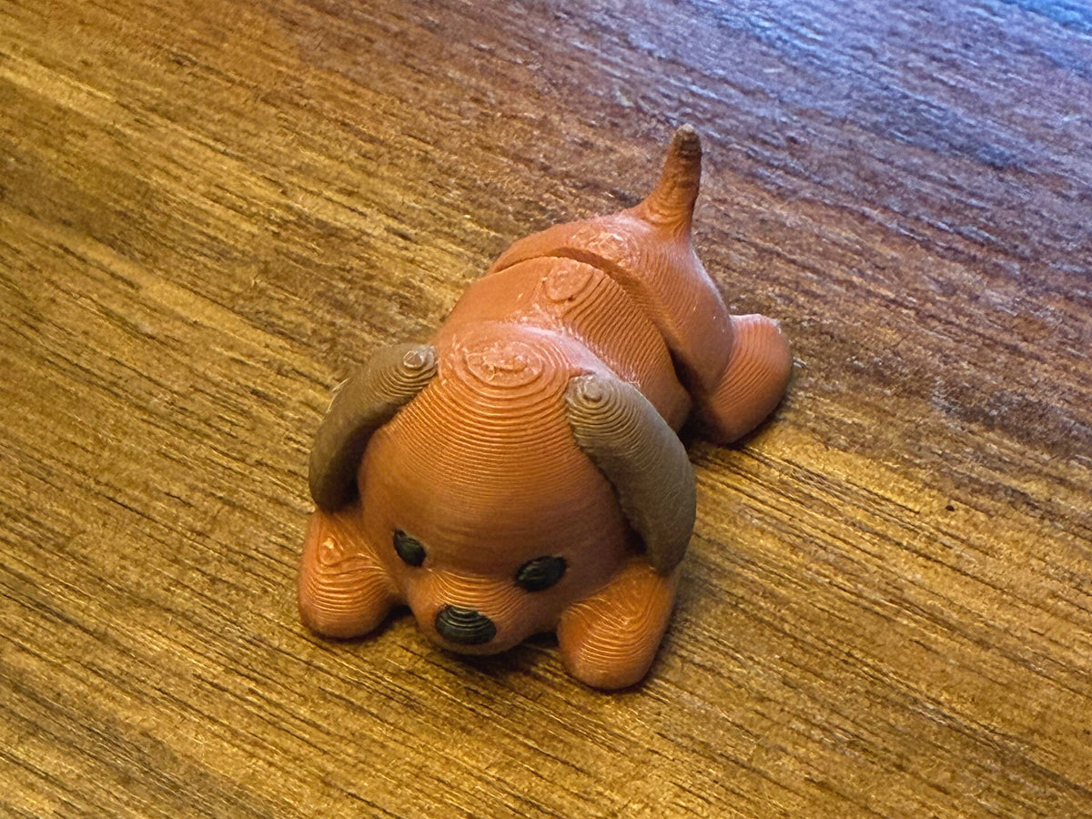 Brown 3D printed Mini Flexi Dog puppy with floppy ears and a black nose, three-quarter front view on a wood table