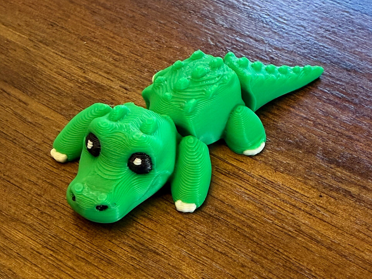 Green 3D printed Mini Flexi Alligator fidget toy with black eyes and white claws, three-quarter view on a wood table