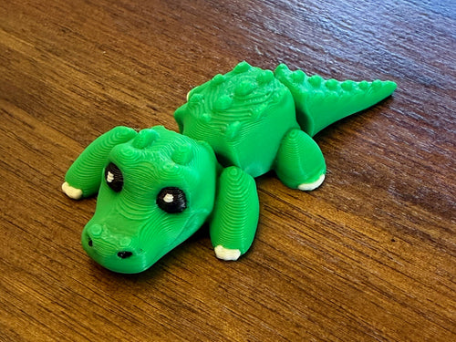 Green 3D printed Mini Flexi Alligator fidget toy with black eyes and white claws, three-quarter view on a wood table