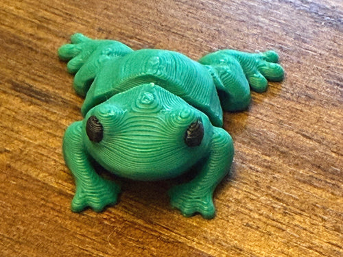 Green 3D printed Mini Flexi Frog with black eyes and webbed feet, three-quarter front view on a wood table