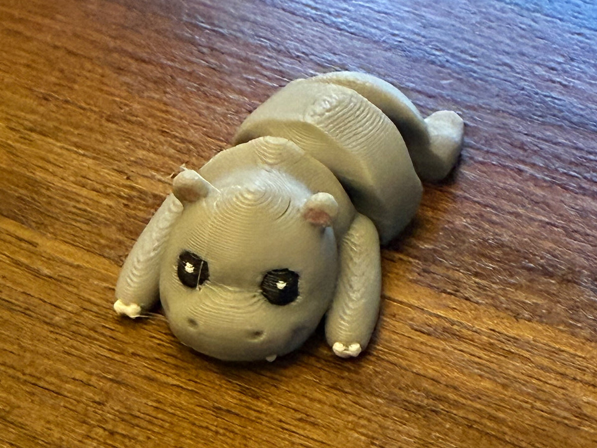 Grey 3D printed Mini Flexi Hippo with pink ears, white toes, and big eyes on a wood table