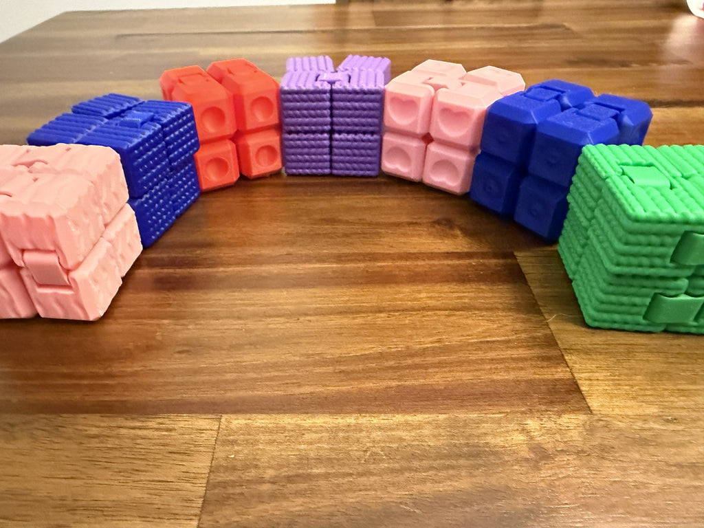 Assortment of 3D printed Infinity Cube fidget toys in pink, blue, red, purple, green, and other colors arranged on a wood table