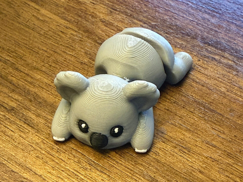 Grey 3D printed Mini Flexi Koala with a black nose and white paw tips, three-quarter front view on a wood table