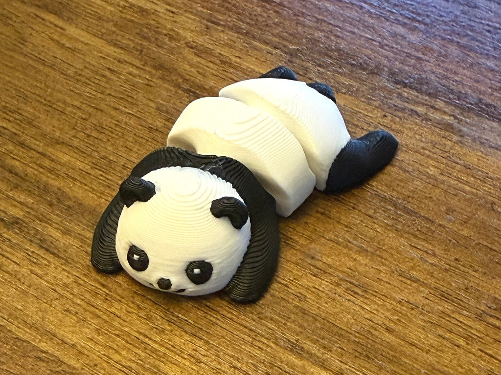 Black and white 3D printed Mini Flexi Panda lying on its belly, three-quarter close-up on a wood table