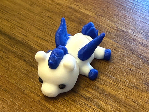 White 3D printed Mini Pegasus figurine with blue wings, mane, and tail, three-quarter view on a wood table
