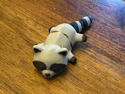 Grey 3D printed Mini Flexi Raccoon with a black bandit mask and ringed tail, three-quarter front view on a wood table